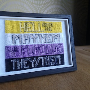 May include: Framed cross-stitch art with a black frame. The artwork features the text "HELL HATH NO MAYHEM LIKE FURIOUS THEY/THEM" in black thread on yellow, white, purple, and black backgrounds. The piece is displayed on a wooden surface.