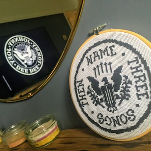 Könnte beinhalten: Ein weißer Stickrahmen mit schwarzen Stichen. Der Rahmen zeigt den Text "NAME THREE SONGS THEN..." mit einem Adlermotiv in der Mitte.
