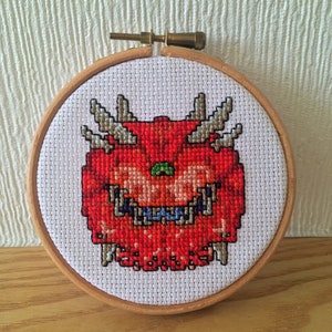 Puede incluir: Un bordado de punto de cruz de una cara de demonio roja pixelada con cuernos grises y una boca azul, en un bastidor de bordado de madera. El diseño está sobre tela blanca y el bastidor se asegura con un tornillo de latón.