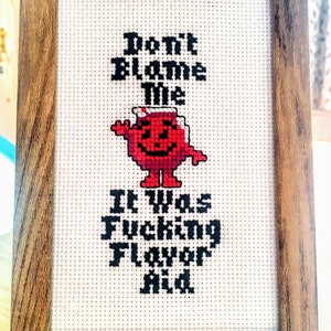 May include: Cross-stitch art in a wooden frame. The design features the text "Don't Blame The" above a red cartoon character, and "It Was Fucking Flavor Aid" below. The frame has a natural wood grain.