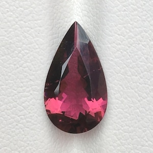 Puede incluir: Una piedra preciosa facetada en forma de pera con un degradado de color magenta intenso y morado oscuro. La piedra presenta una parte superior puntiaguda y una base redondeada con múltiples facetas geométricas que reflejan la luz.