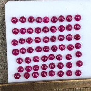 May include: A collection of fifty-two small, round, deep pink cabochon gemstones arranged in rows on a white rectangular surface, positioned next to a brass ruler that shows the stones measure approximately 0.25 inches in diameter each.