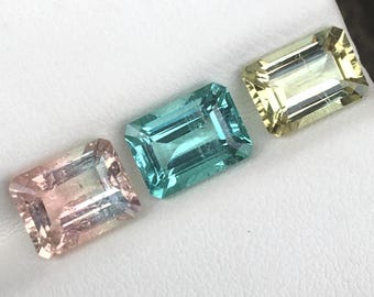 May include: Three rectangular faceted gemstones arranged in a row. The left stone is a pale peach colour, the centre stone is a vibrant teal blue, and the right stone is a light lemon yellow. Each gemstone features a classic emerald cut design.