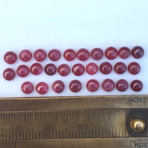 Könnte beinhalten: Eine Sammlung von siebenundzwanzig kleinen, runden, dunkelrosa Cabochons, die in drei Reihen angeordnet sind. Ein Messinglineal liegt unten und zeigt, dass die Steine jeweils einen Durchmesser von etwa 0,6 cm haben.