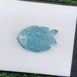 May include: A translucent light blue gemstone carved into the shape of a fish. The carving features detailed scale patterns across the body, a defined head, and a segmented tail fin, showcasing a smooth, polished surface with a glassy appearance.