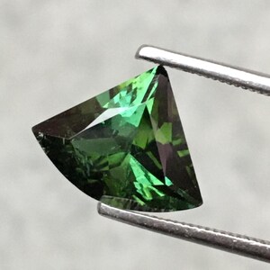 May include: A green gemstone with a triangular shape, held by tweezers. The gemstone has a faceted cut and a dark green color.