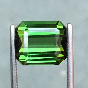 May include: A vibrant green emerald cut gemstone, held in tweezers, with a faceted surface.