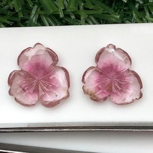 May include: Two carved pink gemstone flower pendants. Each pendant has three petals with etched details and a darker pink center. The gemstones are set against a white surface with green foliage in the background.