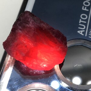 May include: A raw, deep red gemstone with a glowing interior. The stone has a dark, textured exterior and a bright, translucent core. The gem is displayed on a reflective surface, highlighting its natural beauty.