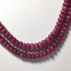 Può includere: Una collana a doppio filo composta da perline lisce, rotonde e a forma di disco di un colore rosso magenta intenso. Le perline sono infilate in un motivo uniforme, creando un design circolare continuo che mostra una consistenza lucida.