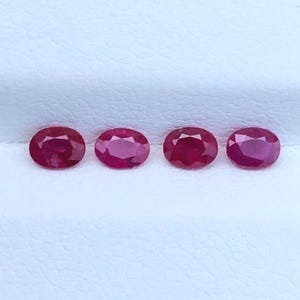 Puede incluir: Cuatro rubíes pequeños de color rojo intenso, con corte ovalado, están dispuestos en una fila horizontal. Cada piedra preciosa presenta una superficie facetada que refleja la luz, mostrando un tono carmesí vibrante sobre una superficie blanca.