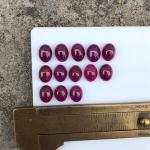 Könnte beinhalten: Dreizehn ovale, tief magentafarbene Cabochon-Edelsteine, die in drei Reihen auf einer weißen Oberfläche angeordnet sind. Ein Messschieber aus Messing befindet sich am unteren Rand und zeigt, dass die Steine etwa 1,3 cm groß sind.