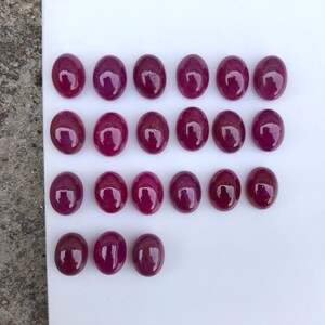 Op de afbeelding: Een verzameling van eenentwintig ovaalvormige, diep magenta gekleurde cabochon edelstenen, gerangschikt in vier rijen op een wit oppervlak. Elke steen heeft een glad, gepolijst en reflecterend oppervlak met een consistente koepelvorm.