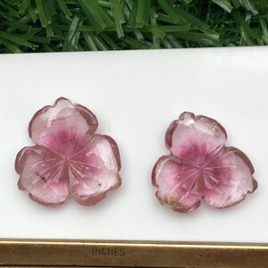 May include: Two carved pink and clear gemstone flower pendants. The pendants are in the shape of a three-petal flower with a darker pink center. A ruler with inch measurements is in the foreground.