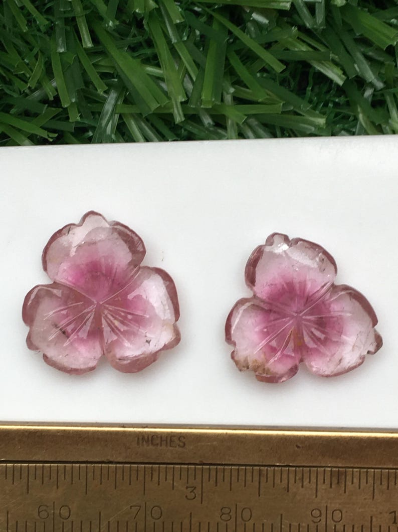 May include: Two carved pink and clear gemstone flower pendants. The pendants are in the shape of a three-petal flower with a darker pink center. A ruler with inch measurements is in the foreground.