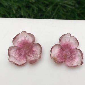 May include: Two pink gemstone flower carvings. Each carving has three petals with etched lines, and a darker pink center. The gemstones are set against a white background, with a blurred green background.
