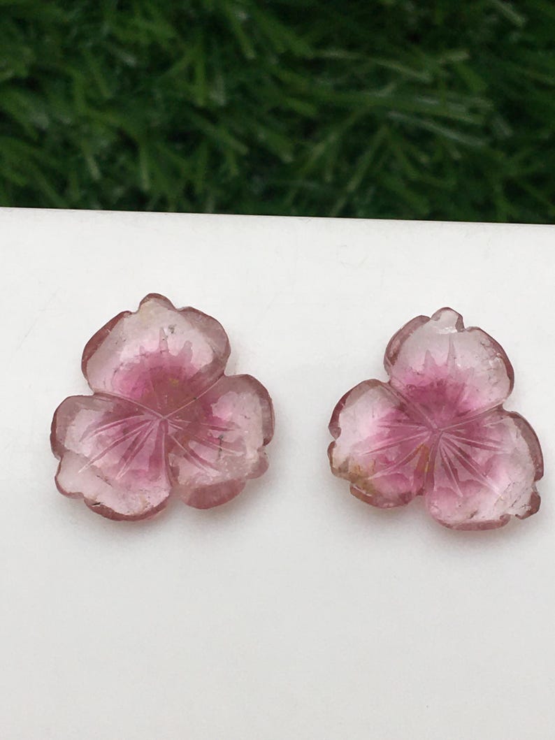 May include: Two pink gemstone flower carvings. Each carving has three petals with etched lines, and a darker pink center. The gemstones are set against a white background, with a blurred green background.