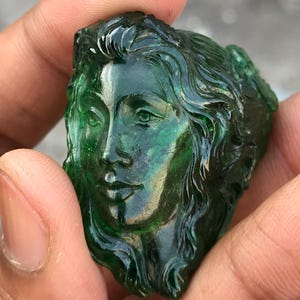 May include: A carved gemstone sculpture of a woman's face. The translucent green stone features detailed facial features and flowing hair. The sculpture is held in a person's hand, showcasing the intricate craftsmanship and the stone's color.