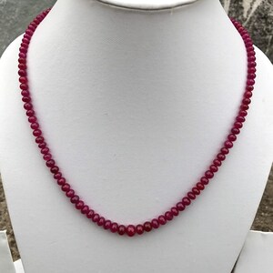 May include: A single strand necklace composed of numerous small, round, deep red beads. The beads are uniform in size and shape, creating a continuous circular pattern that rests against the white display form. The necklace features a consistent color.