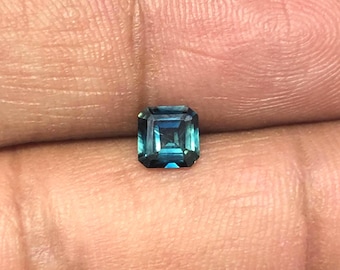 Puede incluir: Una piedra preciosa de corte cuadrado con una paleta de colores azul verdoso profundo. La piedra presenta una superficie facetada que refleja la luz, mostrando una mezcla de tonos azul marino oscuro y cian brillante en su estructura.