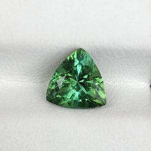 May include: A triangular, green gemstone with multiple facets. The gem's surface reflects light, highlighting its internal structure. The gemstone is set against a white background.