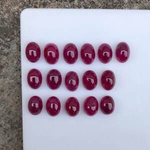 May include: Sixteen oval-shaped, deep magenta-red cabochon gemstones arranged in three rows. Each stone features a smooth, polished, and reflective surface with a consistent rounded dome shape, displaying a uniform saturated colour throughout.