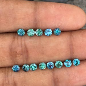 Op de afbeelding: Een verzameling kleine, ronde edelstenen in tinten van teal, turquoise en groen. De edelstenen zijn gerangschikt in twee rijen, waarbij hun verschillende kleuren en interne patronen worden getoond. De stenen worden in een hand gehouden.