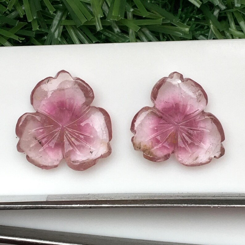 May include: Two carved pink gemstone flower pendants. Each pendant has three petals with etched details and a darker pink center. The gemstones are set against a white surface with green foliage in the background.