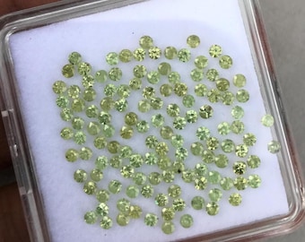 May include: A collection of numerous small, round, faceted lime-green gemstones arranged on a white square display pad inside a clear plastic container. Each individual stone features a brilliant cut with multiple reflective surfaces.