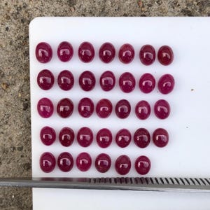 Op de afbeelding: Een verzameling van drieëndertig ovale, dieproze-rode cabochon edelstenen gerangschikt in vijf rijen op een wit oppervlak, met een metalen liniaal aan de onderkant die metingen in centimeters toont voor schaal en referentie.