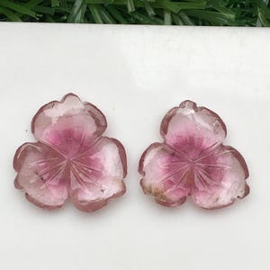May include: Two carved pink gemstone flower pendants. Each pendant features three petals with etched details and a gradient of color from light pink to deep rose. The gemstones are displayed on a white surface.