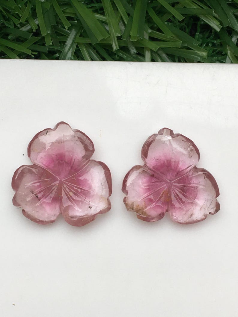 May include: Two carved pink gemstone flower pendants. Each pendant features three petals with etched details and a gradient of color from light pink to deep rose. The gemstones are displayed on a white surface.