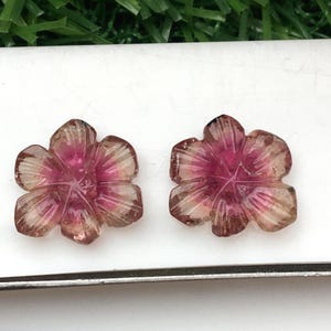 May include: Two carved gemstone flower pendants. Each pendant features a five-petal flower design with a gradient of colors, transitioning from a deep pink center to a translucent, light brown edge. The gemstones are set against a white background.