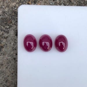 Op de afbeelding: Drie ovaalvormige, diep magenta gekleurde cabochon edelstenen met een glad, gepolijst oppervlak. Elke steen heeft een afgeronde bovenkant en een reflecterende afwerking, met een consistente, verzadigde rood-paarse tint op alle drie.