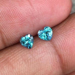 May include: Two heart-shaped, faceted gemstones. The gems are a vibrant teal color, with multiple facets catching the light. The gems are displayed against a neutral background, highlighting their color and shape. These gems could be used for jewelry.