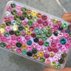 May include: A rectangular clear plastic storage box filled with a large collection of small, round, faceted gemstones in a vibrant array of colours including pink, green, yellow, blue, and red, all arranged on a white base.