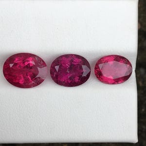 May include: Three oval-cut, deep red gemstones are displayed on a white surface. The gems vary slightly in size and shade, showcasing a range of rich, vibrant hues. The stones are faceted, catching the light and displaying their internal structure.