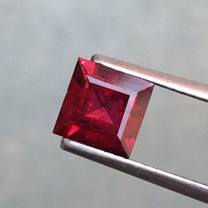 Puede incluir: Una piedra preciosa cuadrada de color rojo oscuro sostenida en unas pinzas. La piedra preciosa está tallada con facetas y tiene un alto brillo.
