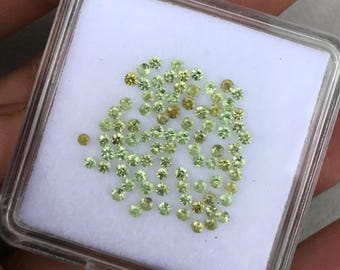 May include: A collection of small, round, faceted gemstones in shades of light green, lime, and pale yellow. These loose, brilliant-cut stones are arranged in a cluster on a white foam insert inside a clear, square plastic display case.