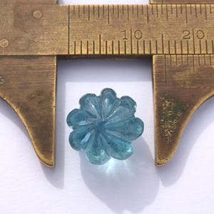 May include: A translucent light blue gemstone bead carved into a flower shape with eight petals. The bead is positioned between the jaws of a brass caliper, showing a diameter of approximately 0.4 inches on the metric scale of the tool.