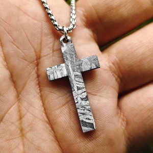 Puede incluir: Un colgante de cruz en tono plateado que presenta un patrón grabado de Widmanstätten en toda su superficie. La cruz está suspendida de una cadena de metal plateado a juego, mostrando un acabado metálico texturizado con líneas geométricas visibles.