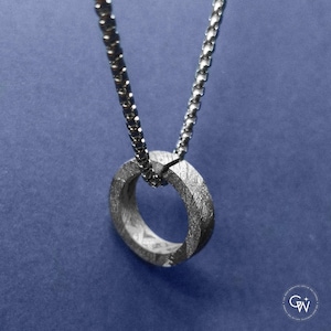 Puede incluir: Un collar con colgante plateado con un dije en forma de anillo texturizado. El dije cuelga de una cadena a juego sobre un fondo azul. El colgante tiene un diseño facetado único.