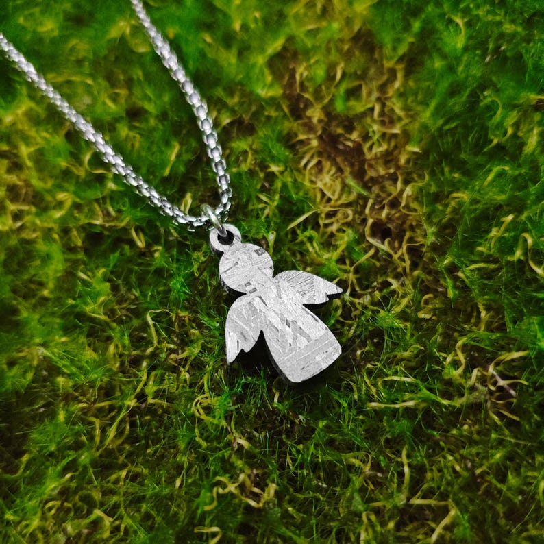 May include: A silver angel pendant necklace with a textured surface. The pendant is on a silver chain and is resting on a bed of green moss.