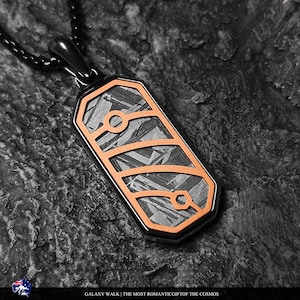 Puede incluir: Un colgante rectangular con marco de metal negro y un patrón geométrico naranja incrustado sobre una superficie metálica oscura y texturizada. El colgante cuelga de una cadena negra e incluye el texto GALAXY WALK | THE MOST ROMANTIC GIFT OF THE COSMOS.