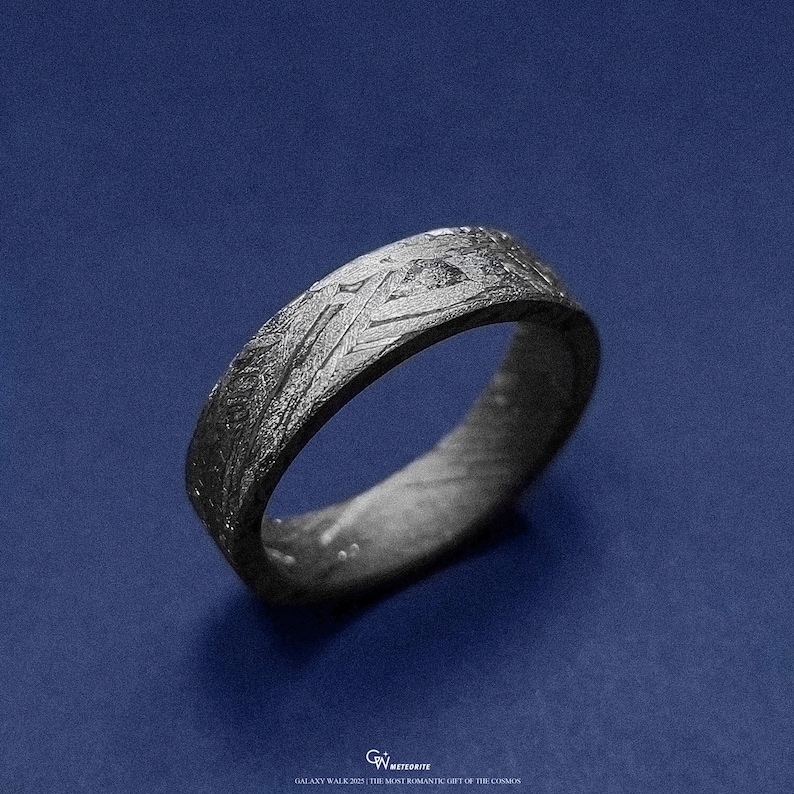 May include: A dark grey ring with a textured surface, set against a blue background. The ring features intricate patterns and designs across its band. The text at the bottom reads "GALAXY WALK 2021 | THE MOST ROMANTIC GIFT OF THE COSMOS."