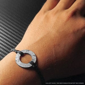 Puede incluir: Una pulsera con un dije circular de color gris metálico con un patrón texturizado y grabado. El dije está sujeto por un cordón negro fino. El texto GALAXY WALK | THE MOST ROMANTIC GIFT OF THE COSMOS aparece en la esquina inferior derecha.