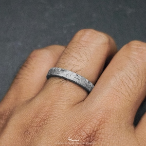 May include: A silver ring with a distinctive textured pattern. The ring is worn on a finger, highlighting its intricate design. The ring's surface has a unique, almost crystalline appearance, possibly indicating a meteorite origin. The ring is a piece of jewellery.