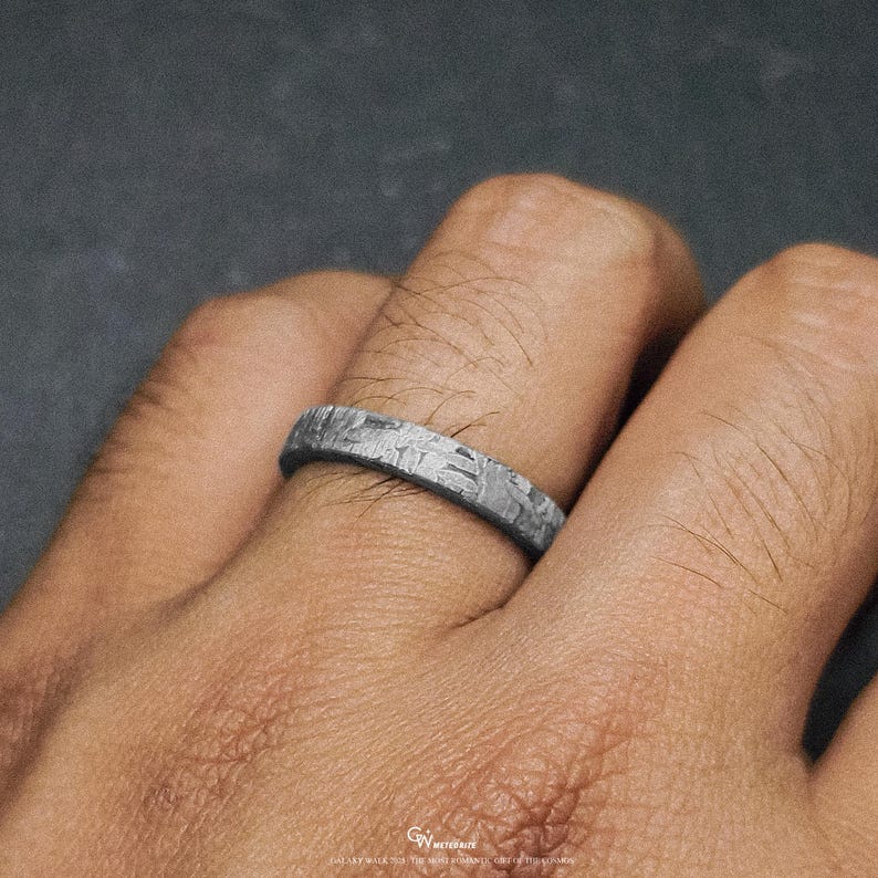 May include: A silver ring with a distinctive textured pattern. The ring is worn on a finger, highlighting its intricate design. The ring's surface has a unique, almost crystalline appearance, possibly indicating a meteorite origin. The ring is a piece of jewellery.