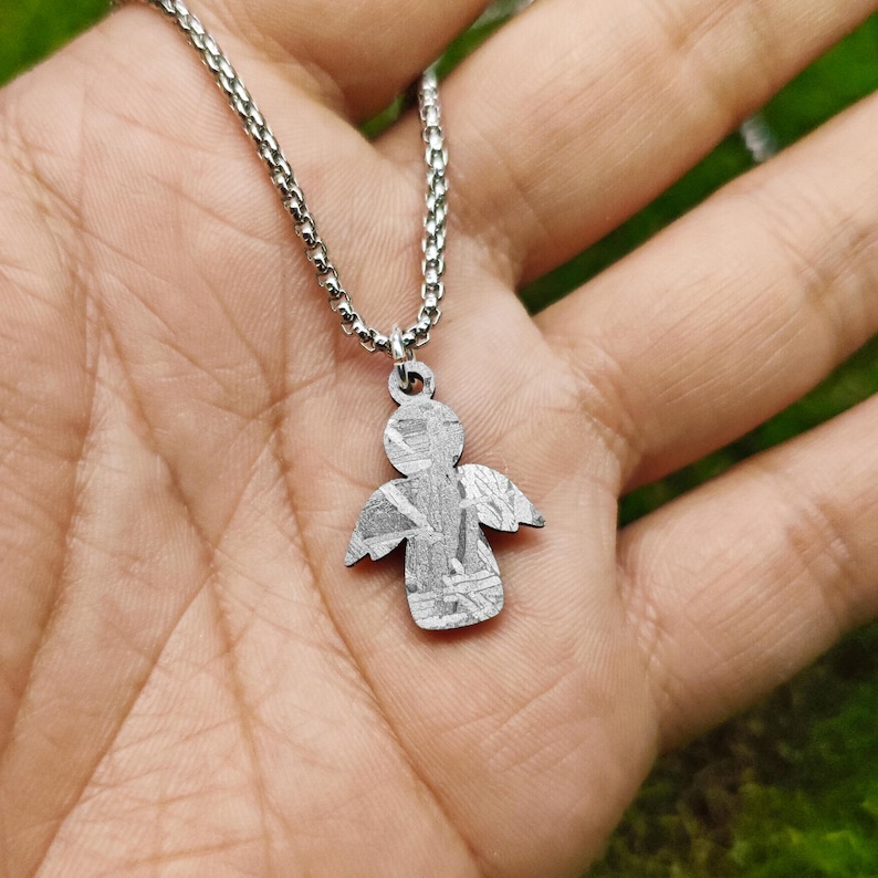 May include: A silver chain necklace with a small angel pendant made of meteorite. The angel has a detailed design with a textured surface.