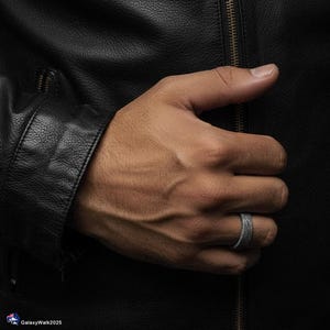 K&ouml;nnte beinhalten: Ein strukturierter grauer Ring wird an einer Hand getragen, vor dem Hintergrund einer schwarzen Lederjacke. Der Ring hat ein bandartiges Design und scheint aus einem matten Material zu bestehen. Das Bild ist gut beleuchtet und hebt die Textur des Rings hervor.